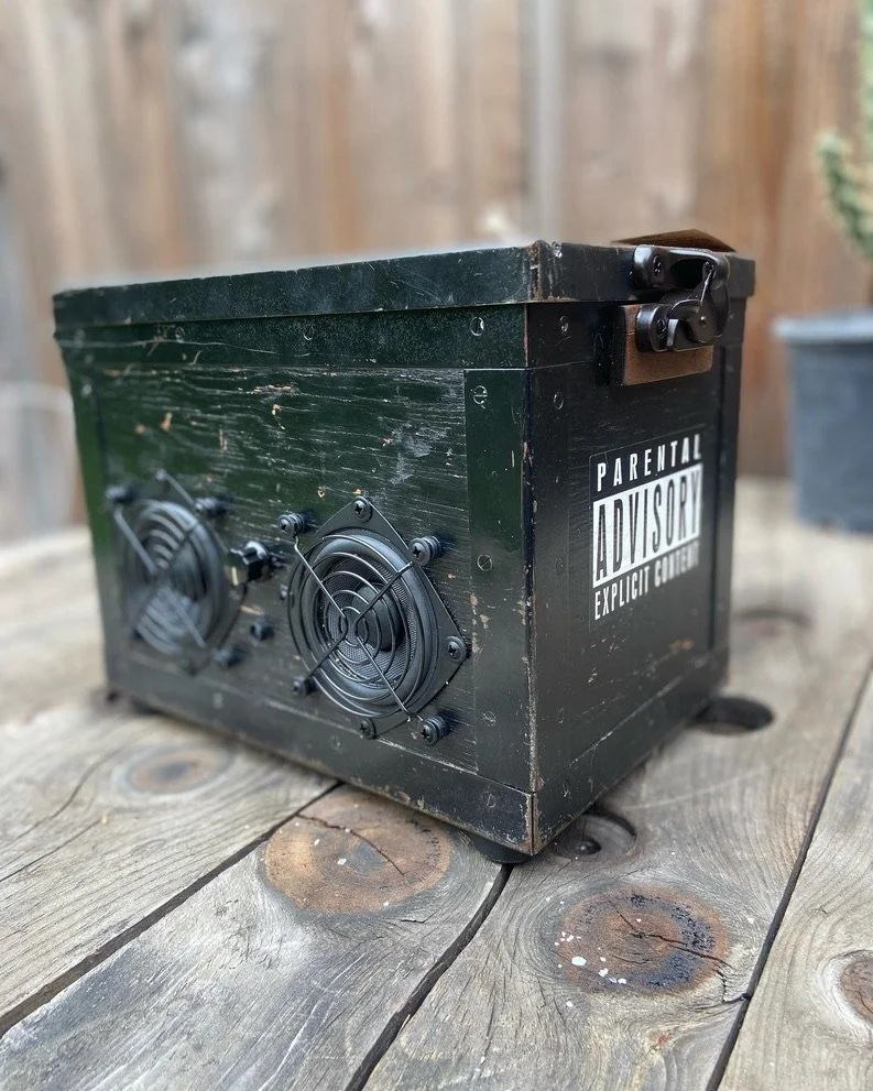 Vintage black wooden box with metal latches and a "Parental Advisory Explicit Content" sticker, equipped with two small speaker fans, sitting on a rustic wooden surface.
