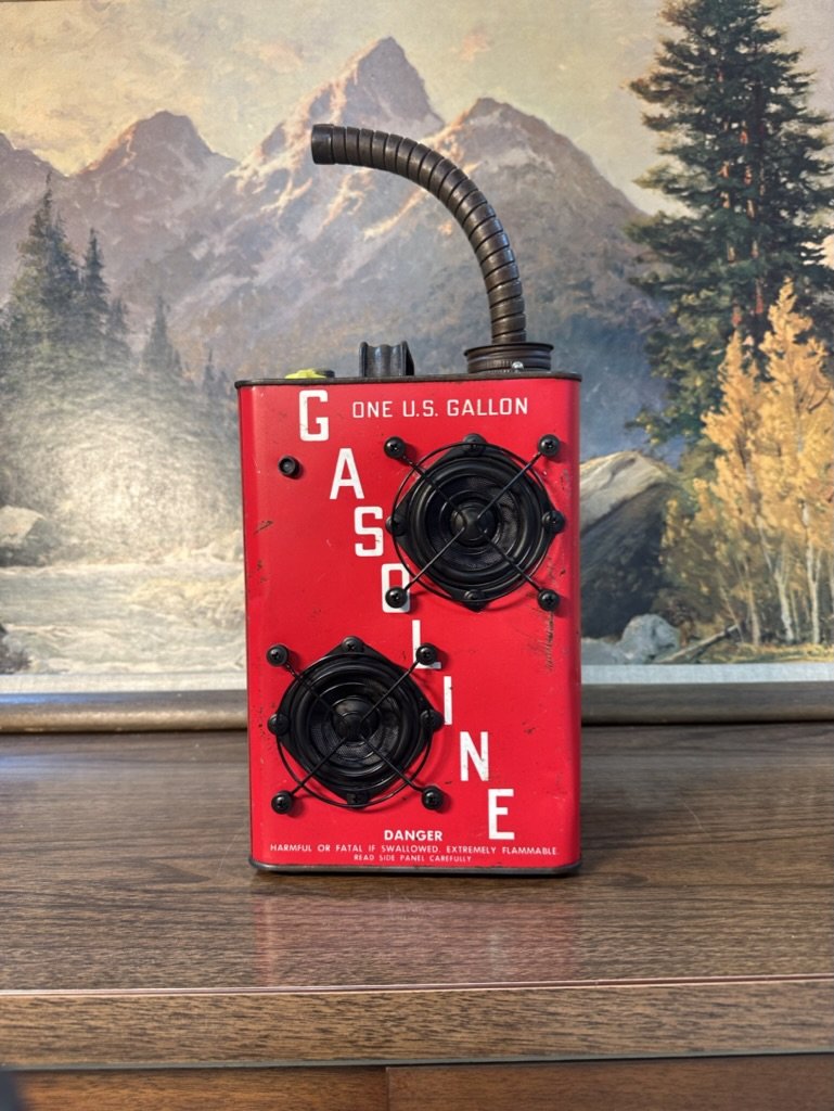 Red vintage one-gallon gasoline can with attached black speakers and flexible conduit on a wooden surface, set against a mountain landscape painting.