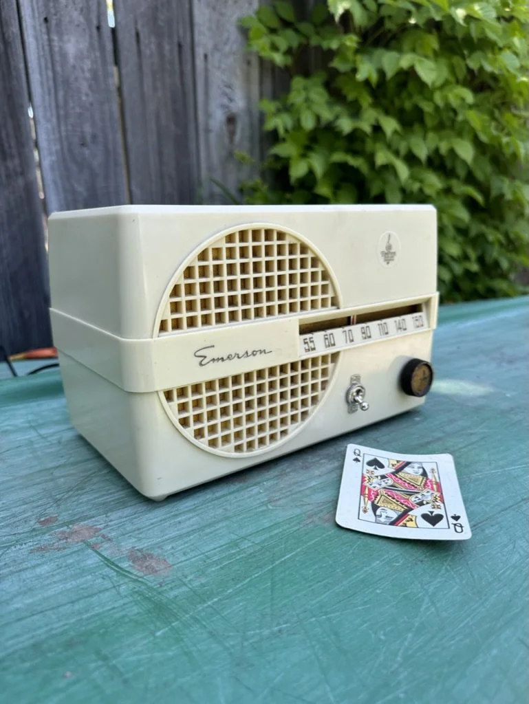 Vintage Emerson radio with a tuning dial and speaker grille, placed on a green surface next to a Queen of Spades playing card, with a wooden fence and greenery in the background.