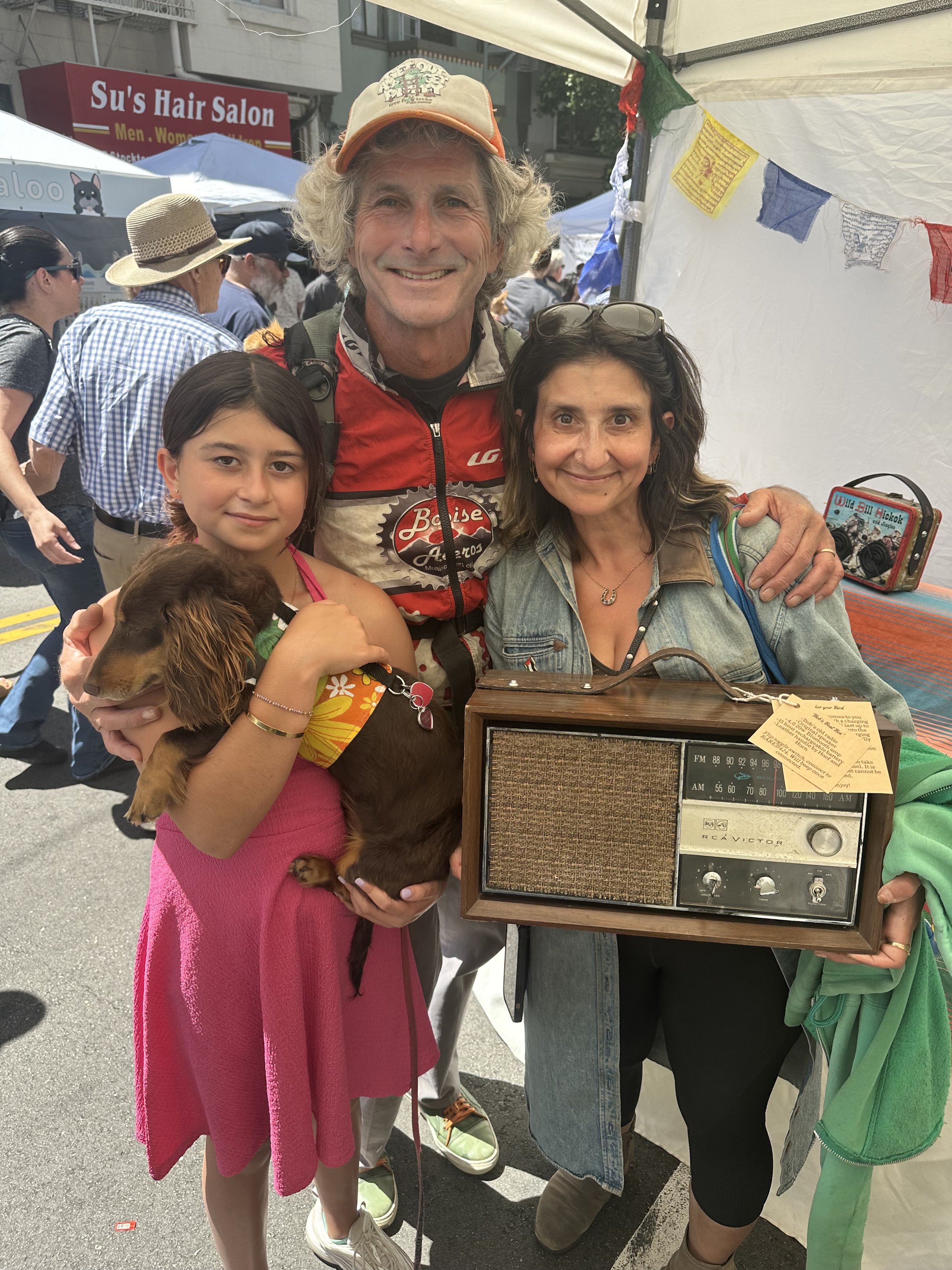 Group of three people at a street fair; a girl in a pink dress holding a dog, a man in a cap with his arm around both, and a woman holding a vintage radio.