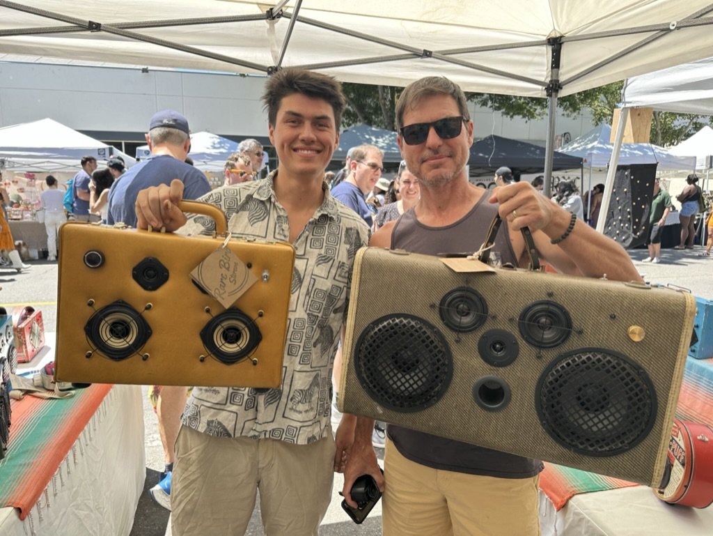 Two men holding handmade suitcase speakers at an outdoor market under a canopy tent, with other market stalls and people in the background.