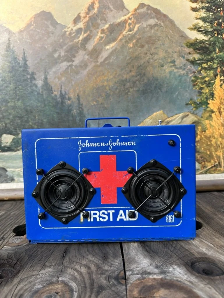 Blue vintage first aid box with speakers attached, placed on a wooden surface, with a mountain landscape painting in the background.