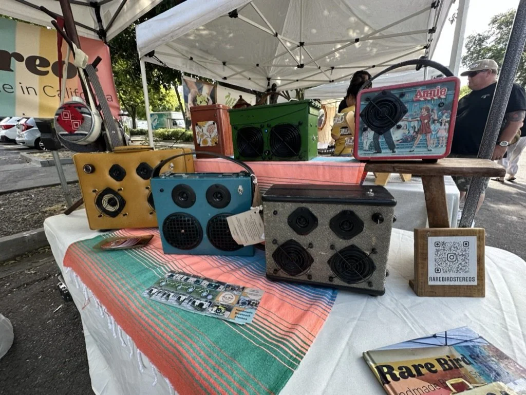 Vintage-style stereos displayed on a table under a white tent at an outdoor market. Items include colorful rectangular cases with speakers, resembling retro radios. A QR code sign with "Rare Bird Stereos" is visible. The table is covered with a strip