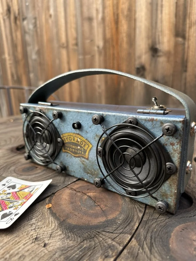 Vintage portable speaker made from a metal box with two visible speakers, a handle on top, and a toggle switch. A playing card is placed beside it on a wooden surface.