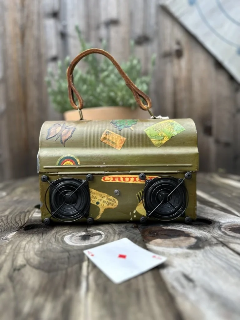 Vintage lunchbox with stickers and built-in speakers on a wooden table, with a playing card (ace of diamonds) lying in front. Potted plant in the background.
