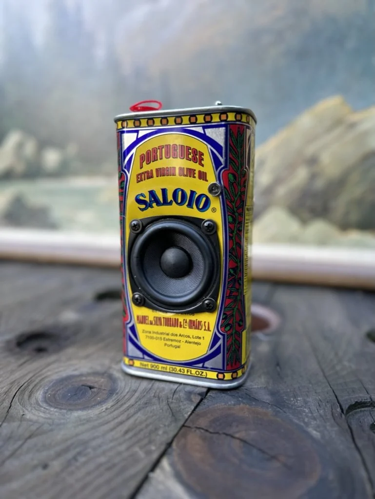 Decorative tin of Saloio Portuguese extra virgin olive oil with a faux speaker design, placed on a wooden surface.