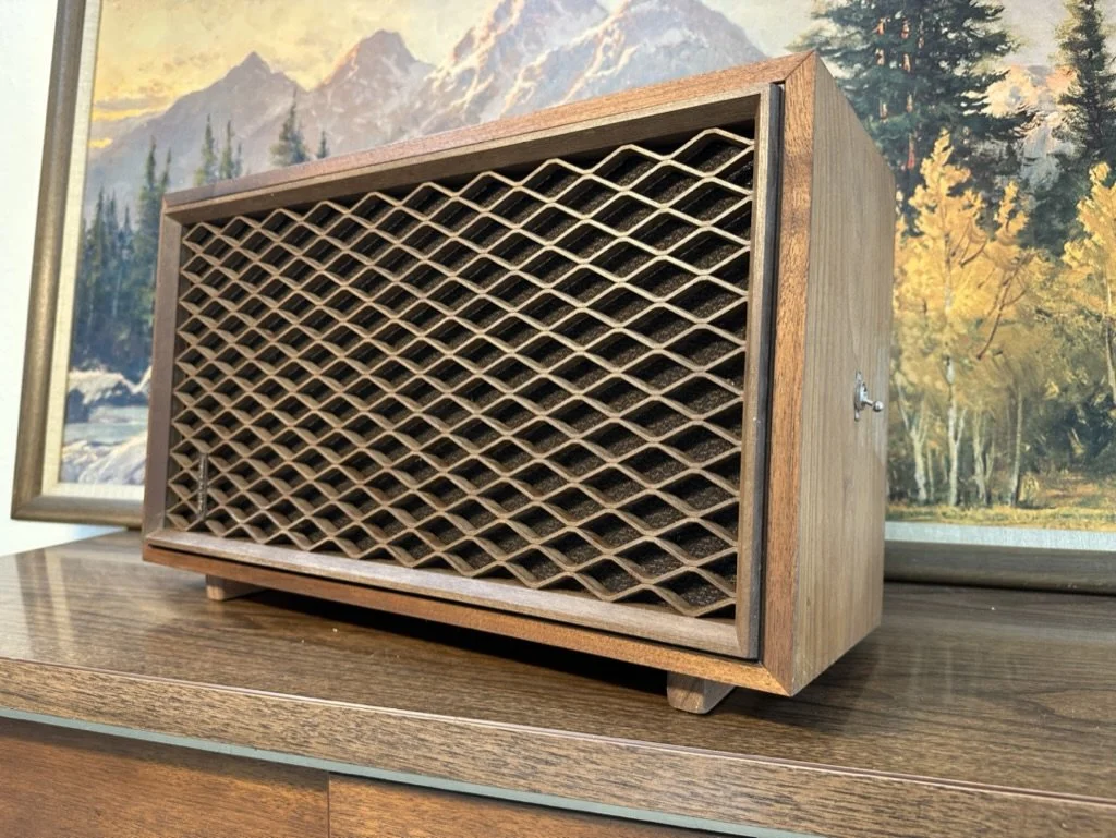 Vintage wooden speaker with lattice grille, standing on a wooden surface, with a landscape painting in the background.