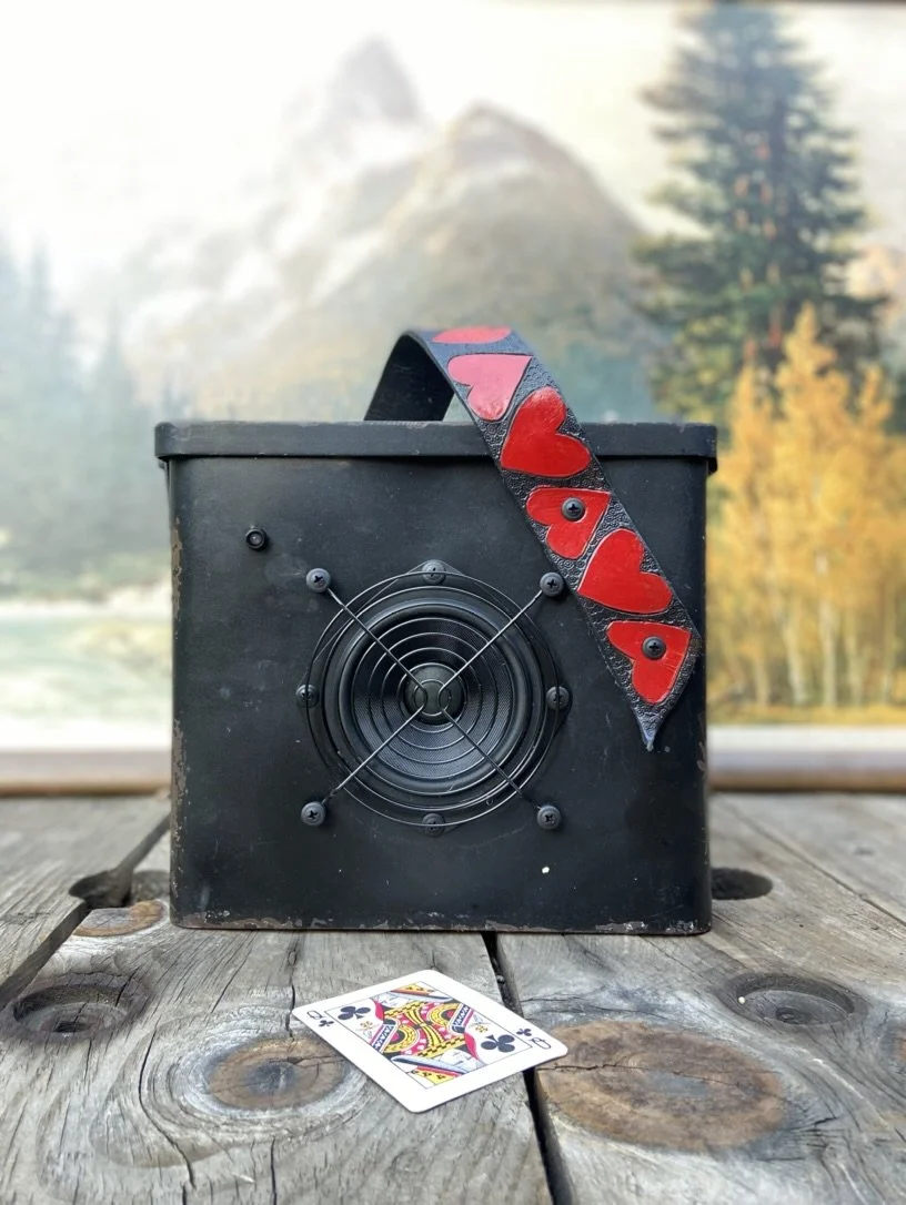 A black box with a fan on one side and a playing card depicting the queen of clubs on a wooden surface. A strap with red heart designs is draped over the box. The background features a blurred outdoor scene with trees and mountains.