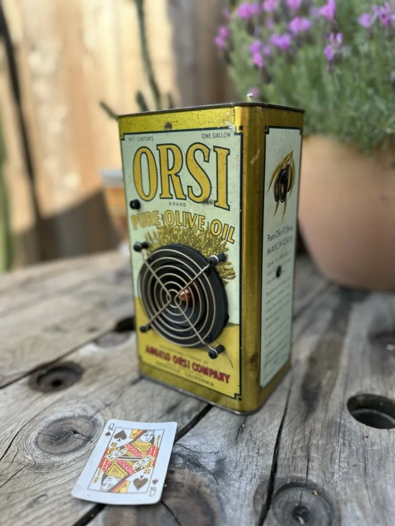Vintage olive oil can labeled "ORSI Pure Olive Oil" with a fan attached, placed on a rustic wooden table next to a Queen of Spades playing card.