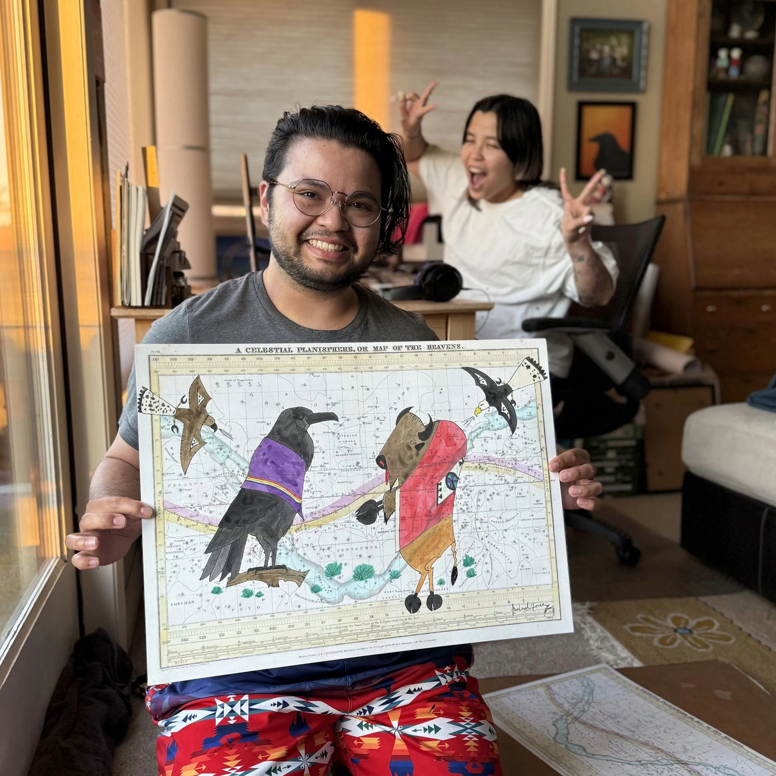 Raven smiles and holds a ledger art drawing of a raven and a bull on a map of the night sky