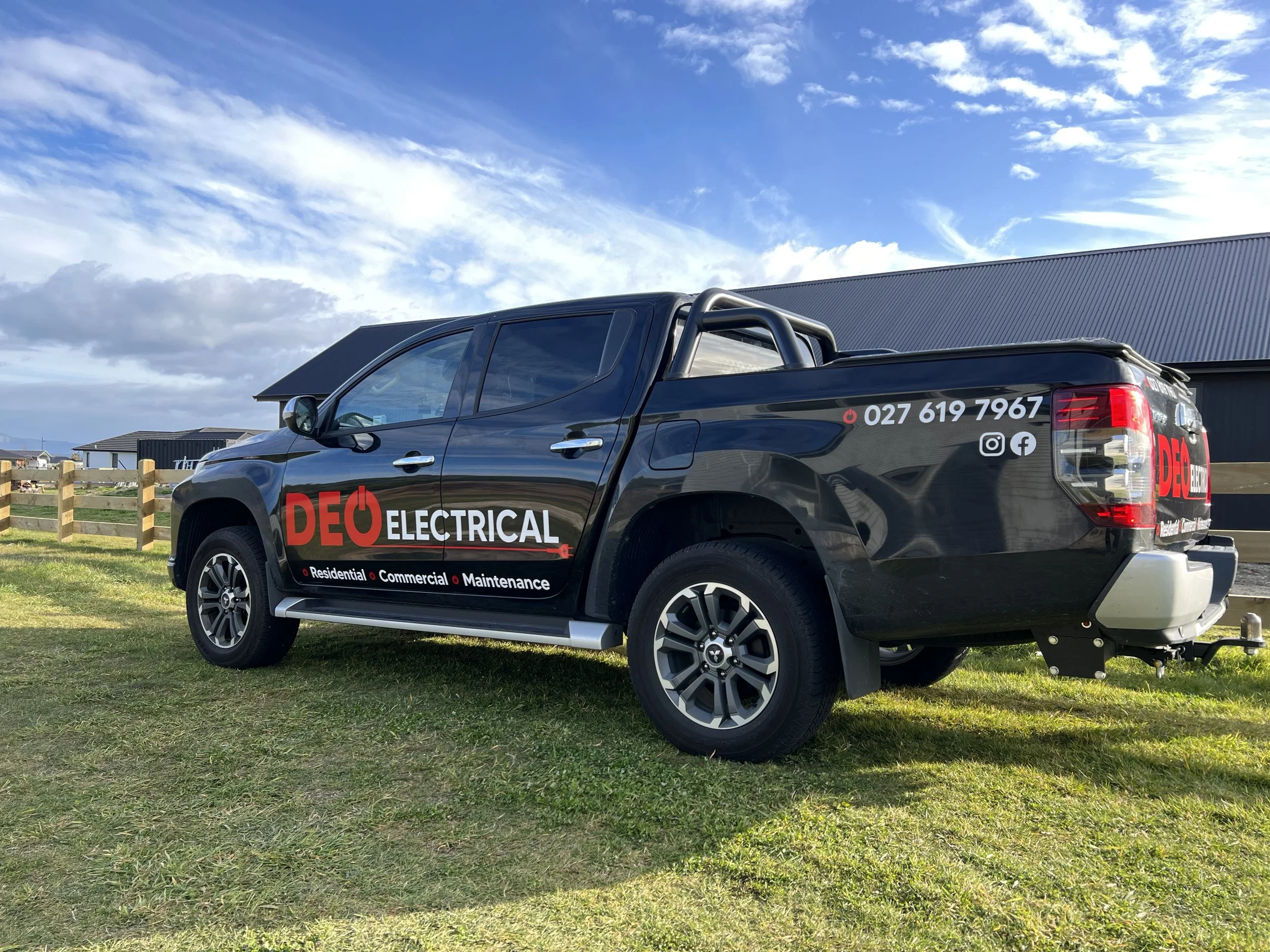 Black pickup truck with 'DEQ Electrical' logo and contact information parked on grass near buildings under partly cloudy sky.