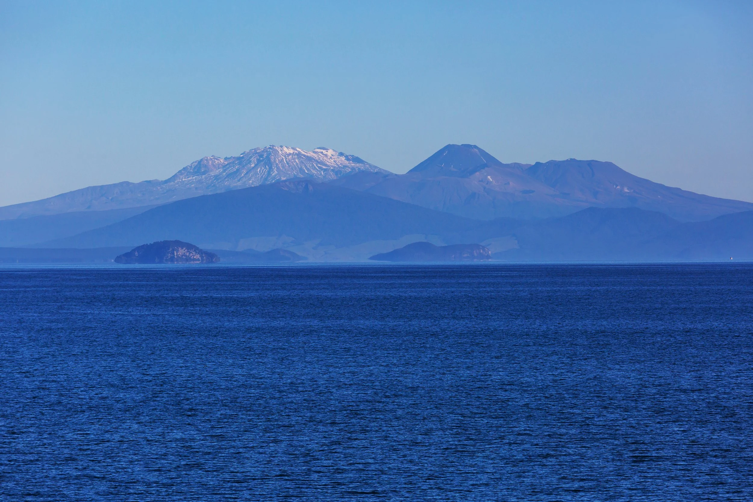 A view of Mount Fuji with snow on its peak, seen across a large body of water, possibly a lake, with small islands and a clear blue sky in the background.