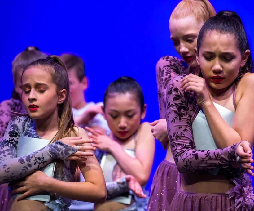 Young dancers on stage in coordinated costumes performing with expressive gestures and closed eyes.