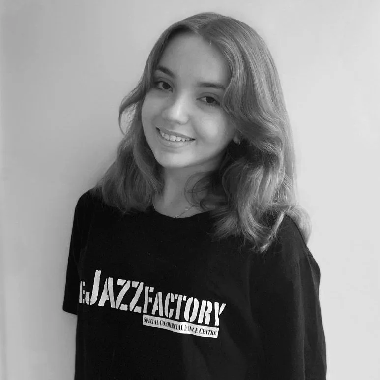 Person wearing a black "Jazz Factory" t-shirt, smiling with shoulder-length hair, posed against a plain background in black and white.