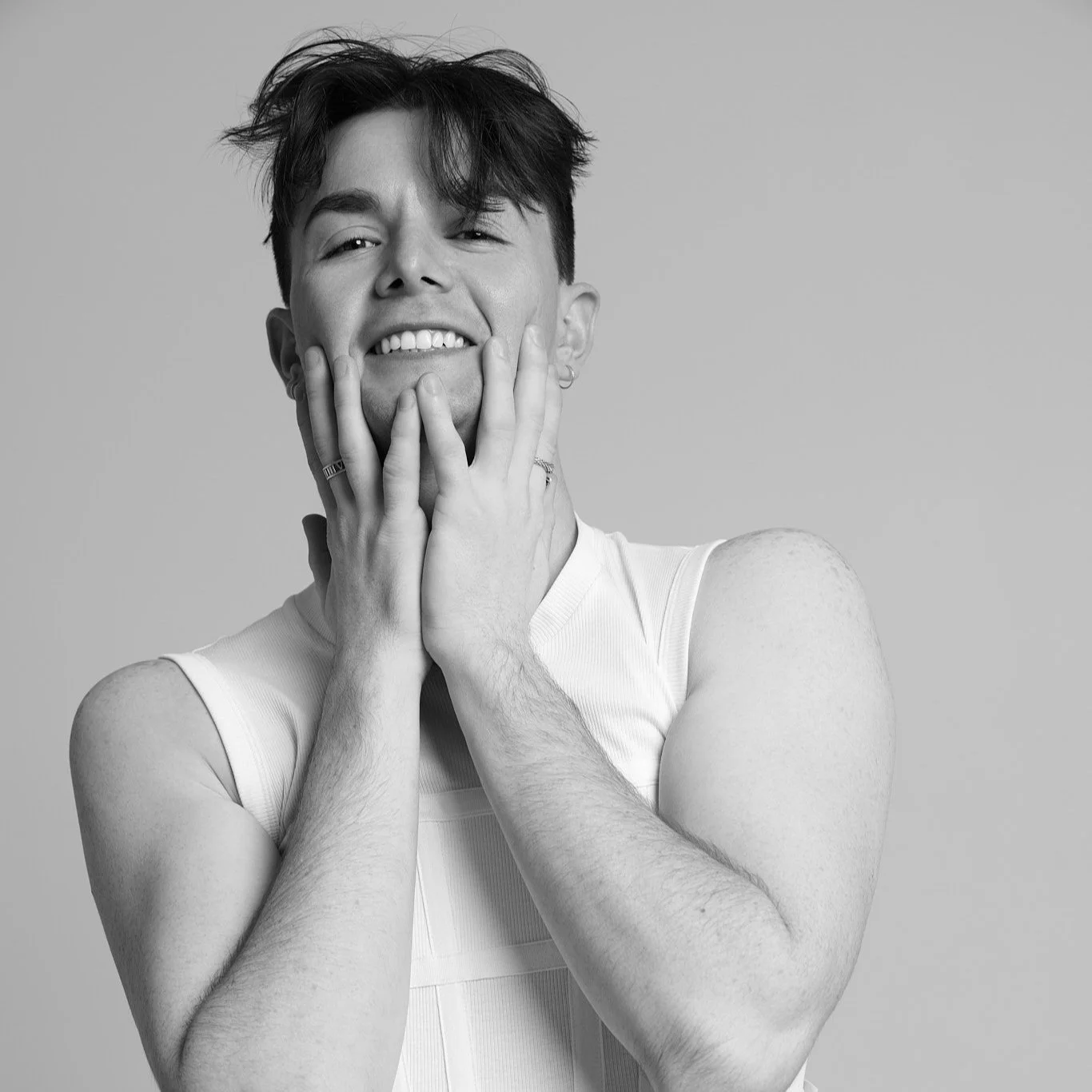 Black and white portrait of a smiling individual with hands on cheeks, wearing a sleeveless top.