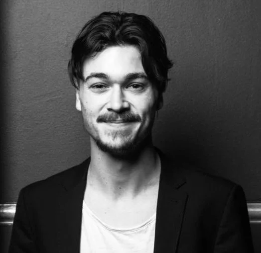A black and white portrait of a man with medium-length hair and a beard, wearing a blazer and t-shirt, in front of a plain background.