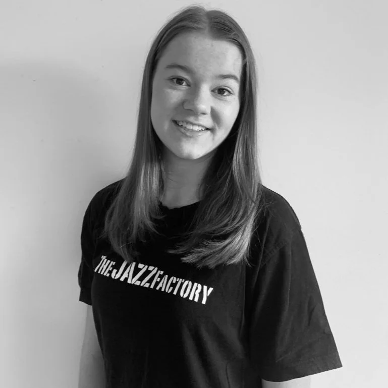 Black and white photo of a smiling individual wearing a t-shirt with 'The Jazz Factory' text on it.