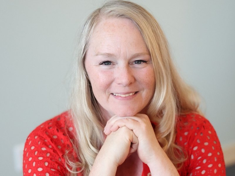 A smiling woman with long blonde hair in a red polka dot shirt, resting her chin on her hands.