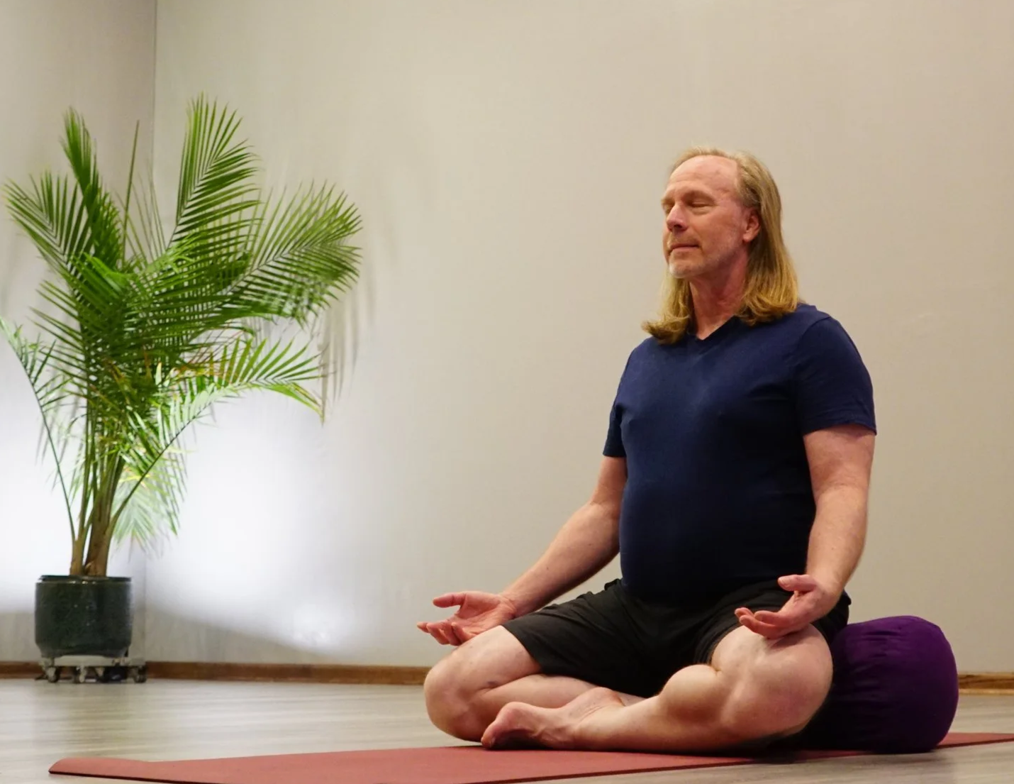 A man with long blond hair, wearing a navy blue shirt and black shorts, practicing meditation on a yoga mat, seated in a cross-legged position, with a calming expression. There is a potted plant with large green leaves in the background.
