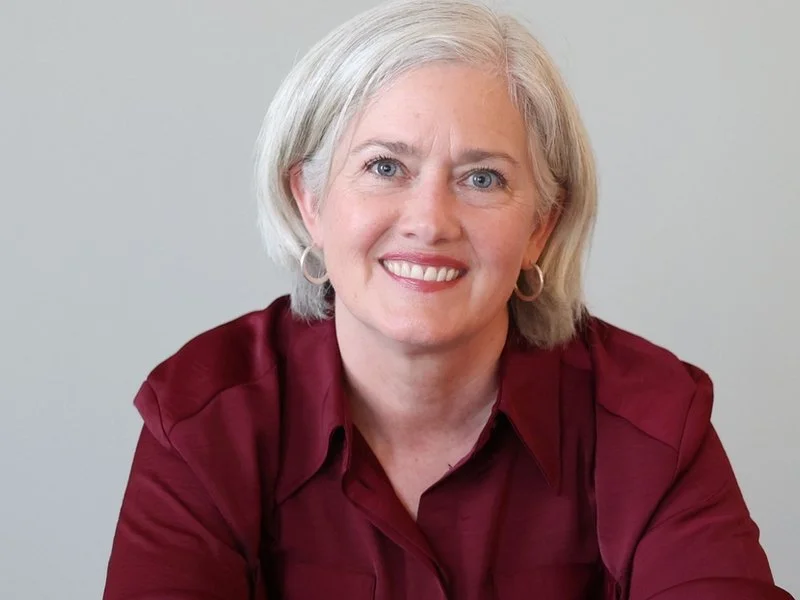 A woman with short, gray hair and blue eyes, wearing a maroon blouse and hoop earrings, sitting and smiling in front of a plain light gray background.