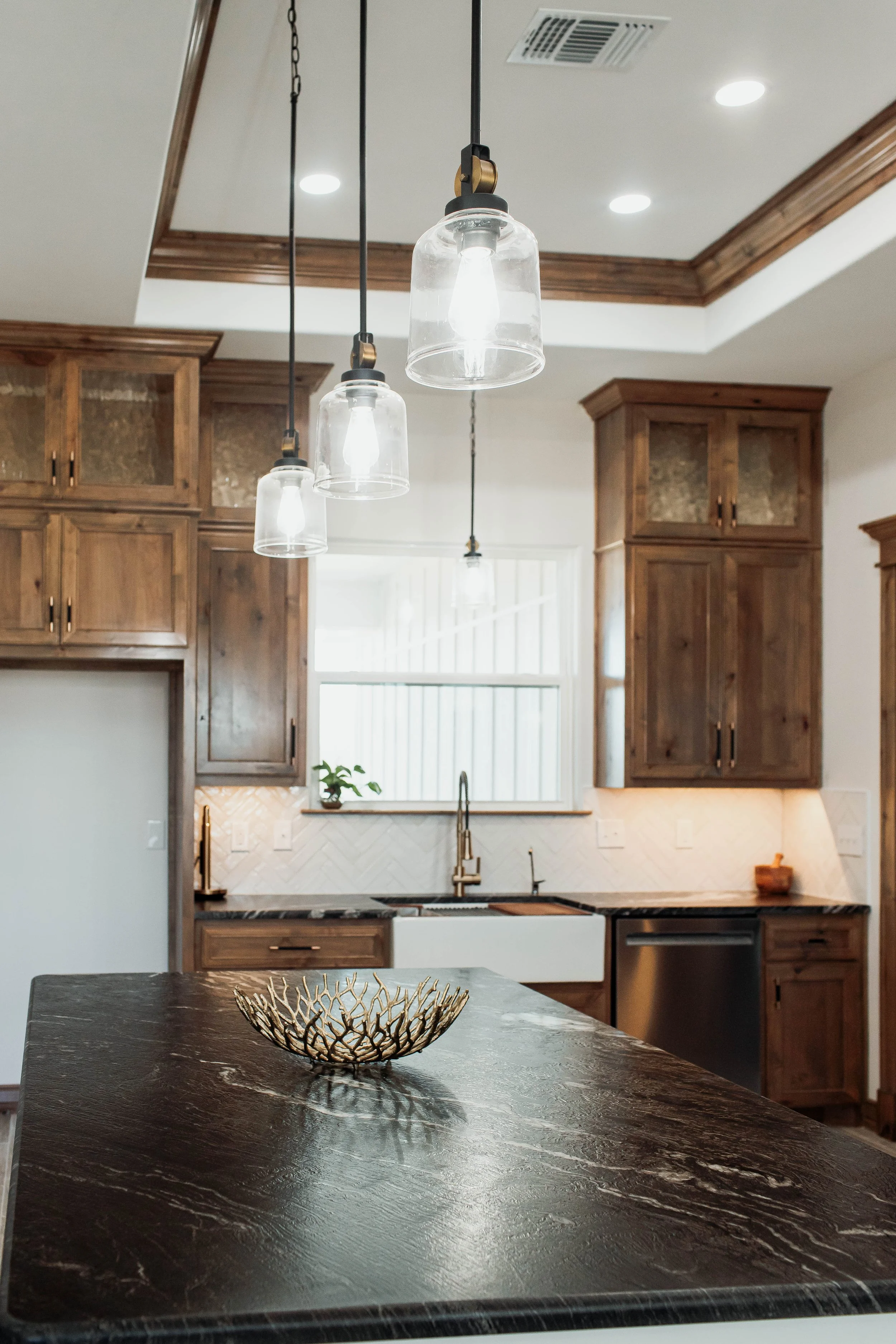 A kitchen with dark wood cabinets, a marble countertop, a window, and a chandelier with three glass shades hanging from black cords.