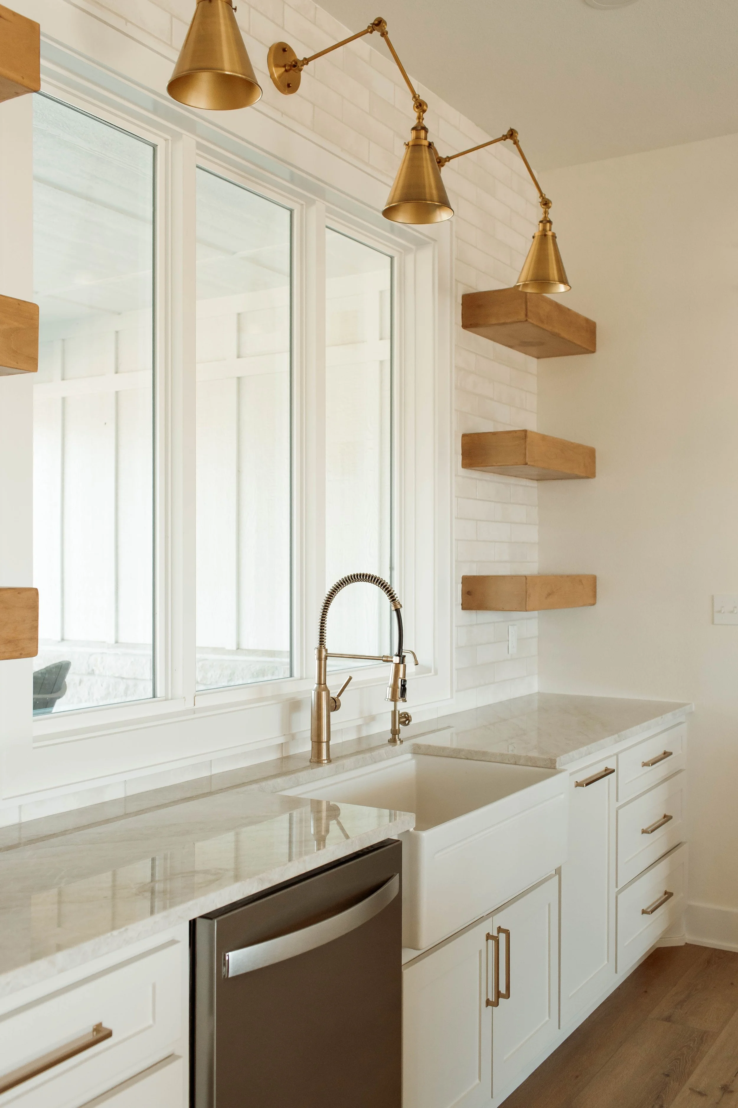 Modern kitchen interior with white cabinets, a marble countertop, a gold-colored faucet, large window with white frames, and three gold wall-mounted lamps on a white brick wall, with wooden shelves.