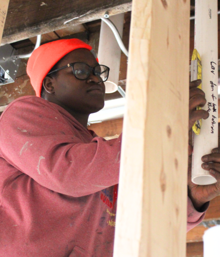 A person wearing a red beanie, glasses, and a dusty pink sweatshirt, working on a construction or plumbing project with a white pipe, in an unfinished building.