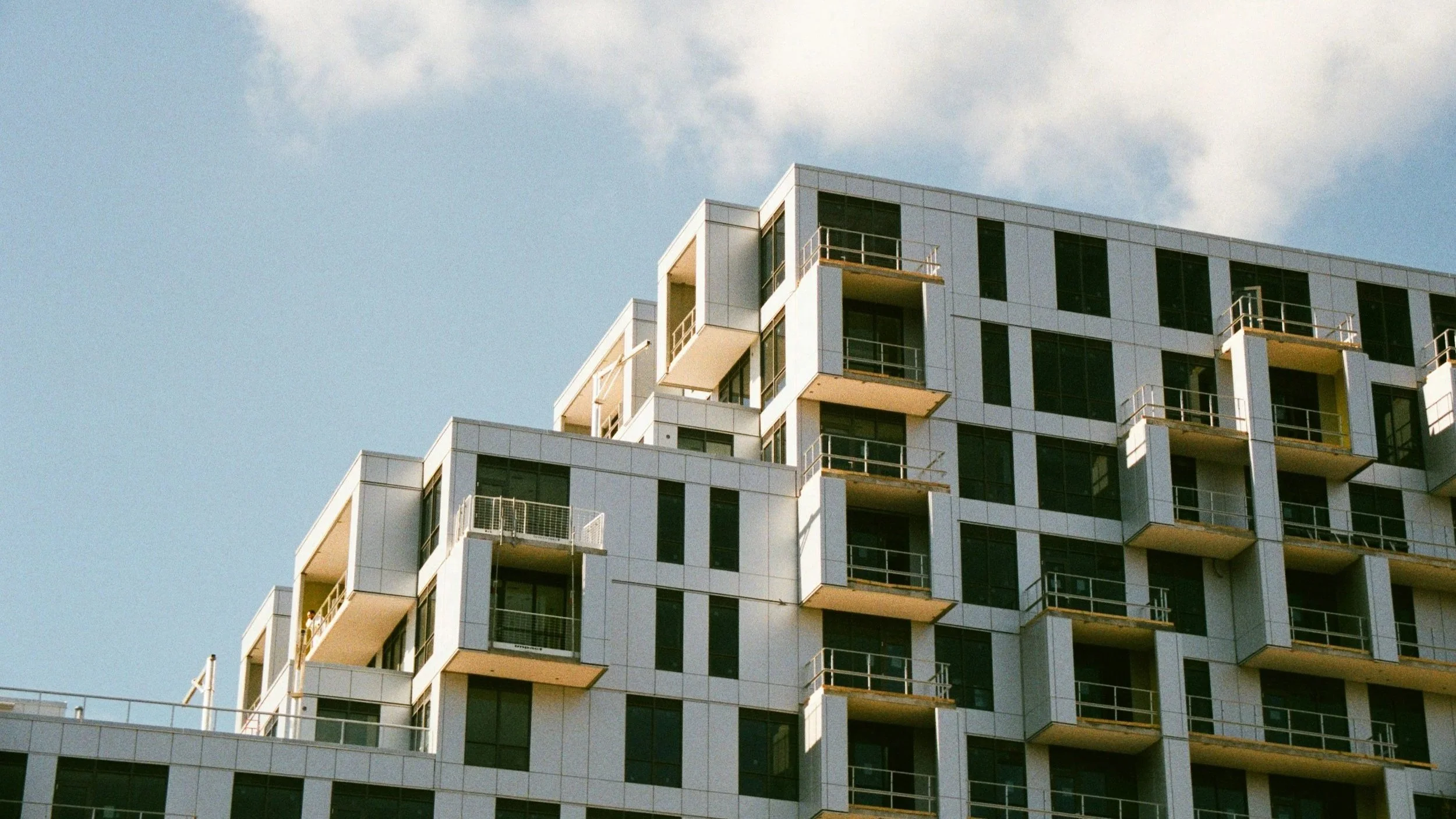Modern multi-story residential building with balconies, large windows, and a white exterior against a blue sky with some clouds.