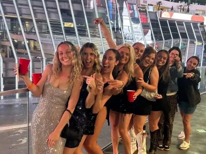 Young women in stylish dresses posing together at a party or nightclub with other people dancing in the background.