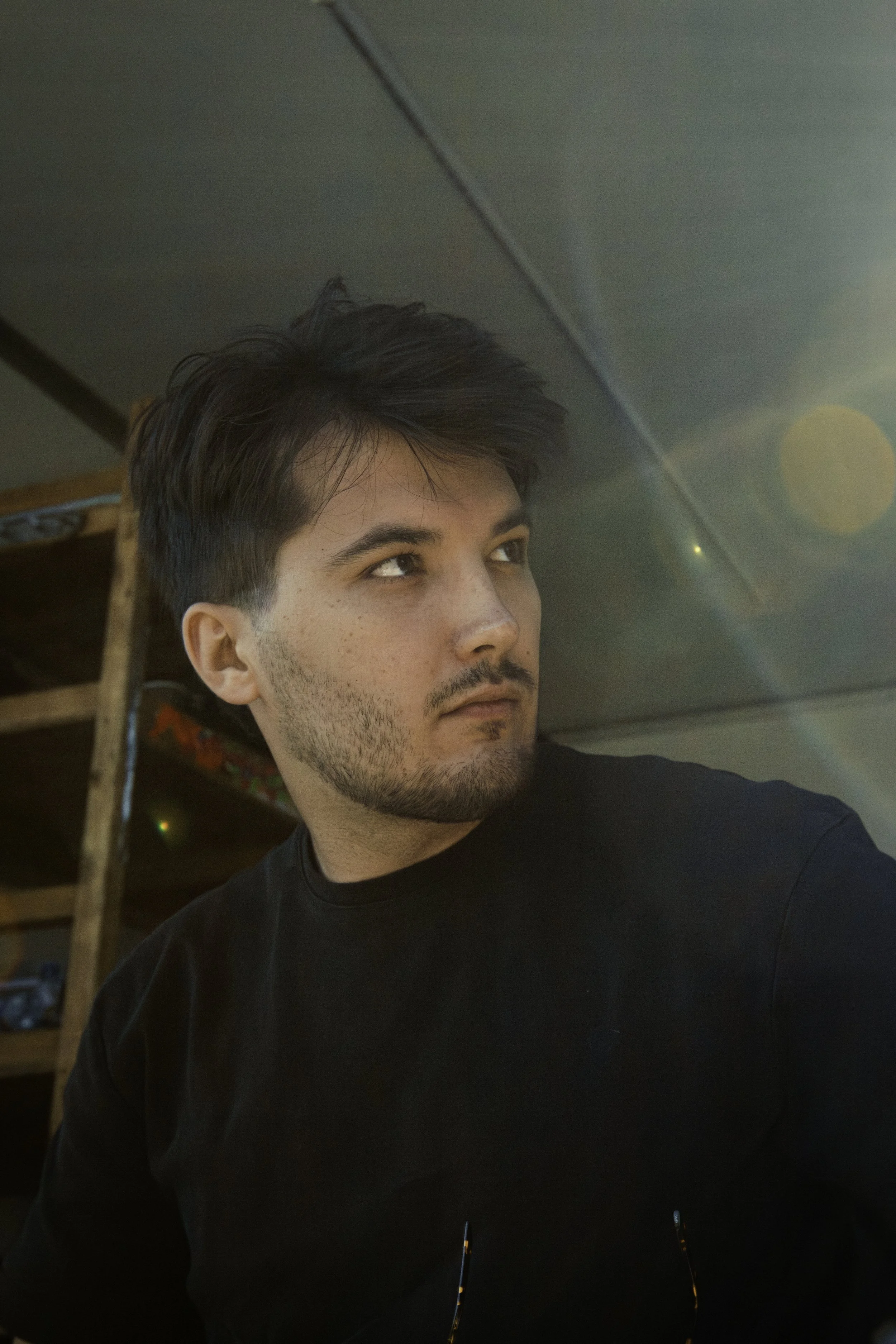 A young man with dark hair and facial hair is looking to his left. He is indoors, wearing a black shirt, with a wooden shelf in the background and sunlight creating lens flare effects.