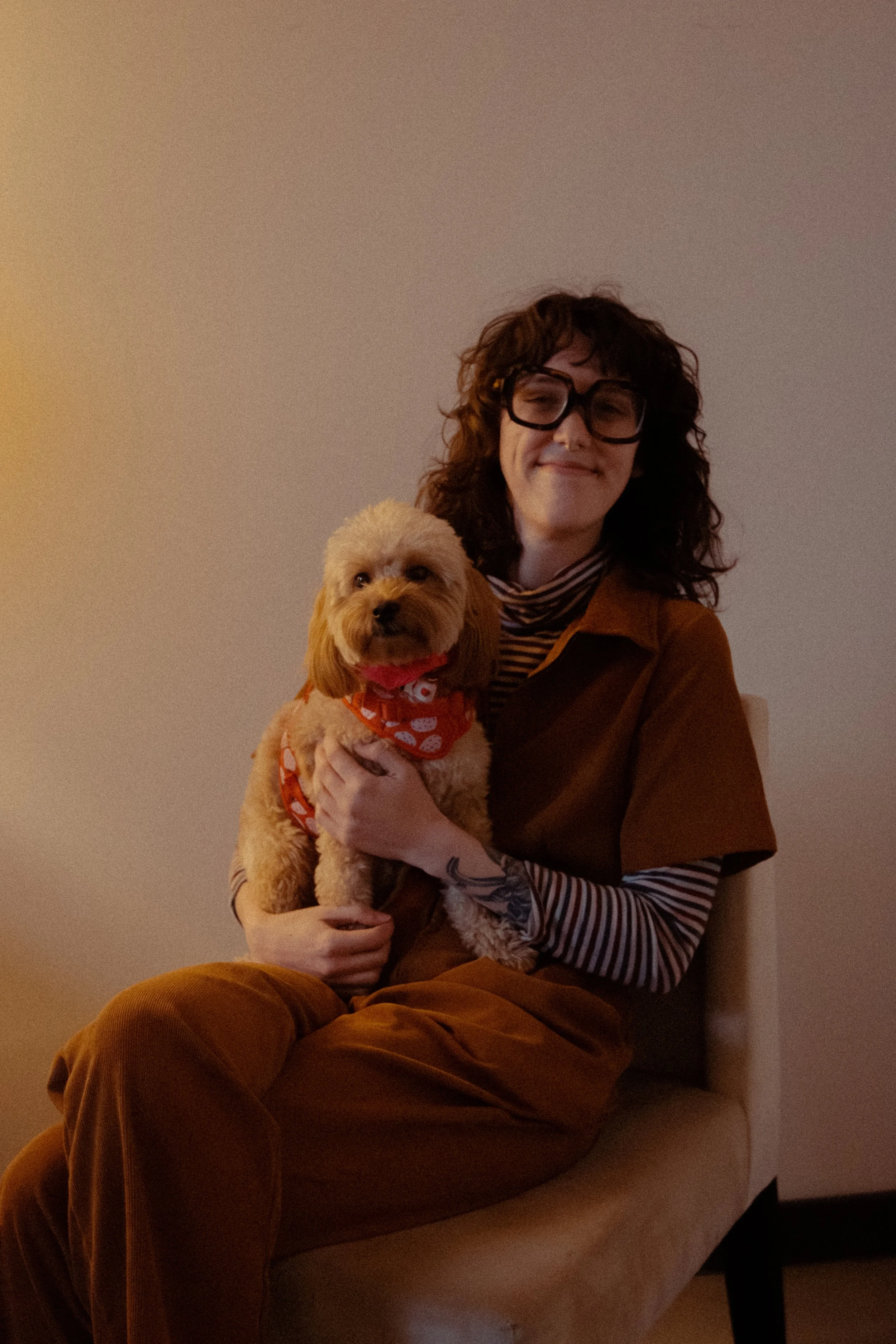 A woman with curly hair, glasses, and wearing a striped shirt and brown clothing, sitting on a chair, holding a small fluffy dog with a red bandana, in a room with plain walls.