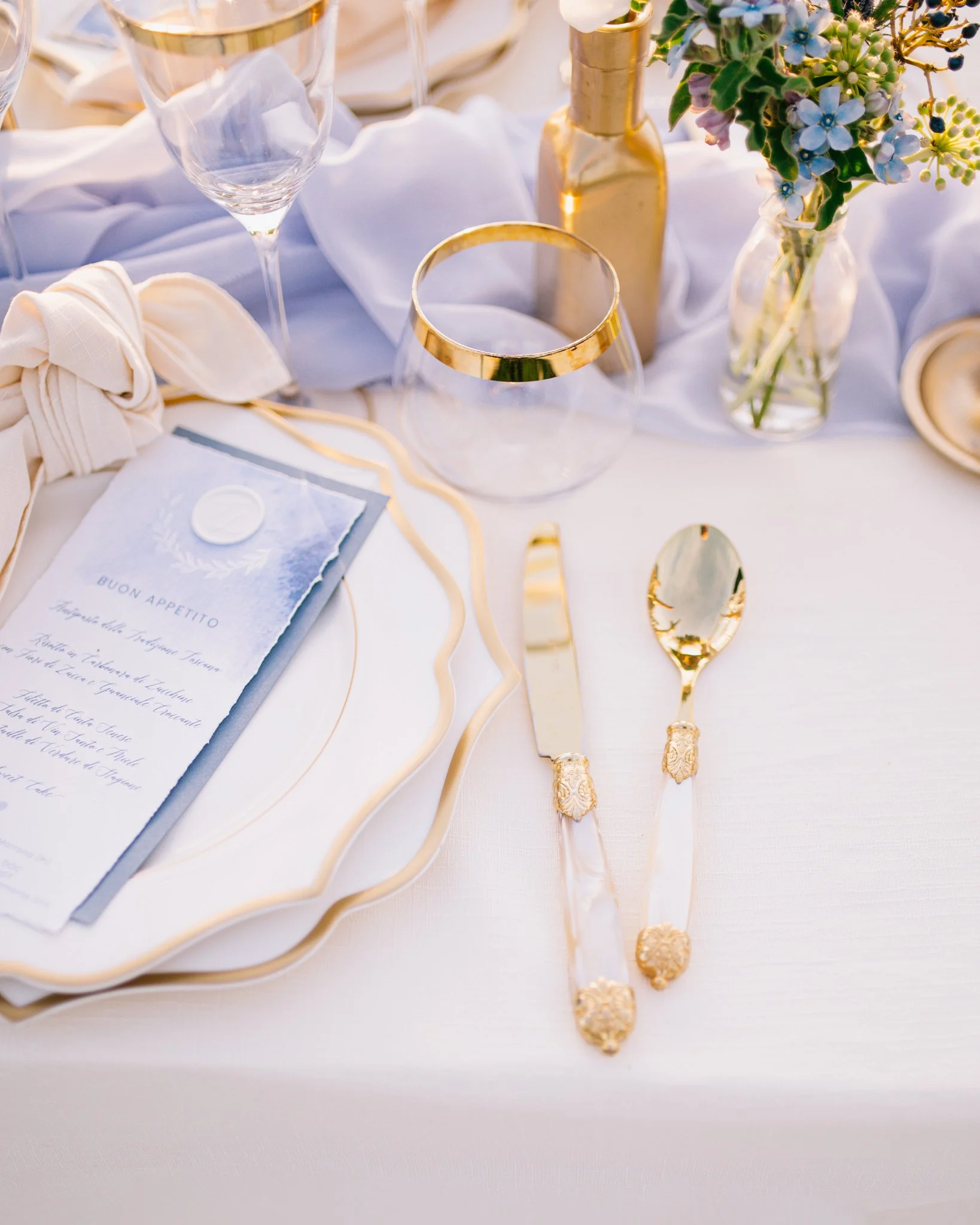 Elegant table setting with gold-accented glassware, a gold vase with blue and green flowers, a menu, and gold-handled cutlery on a white tablecloth.