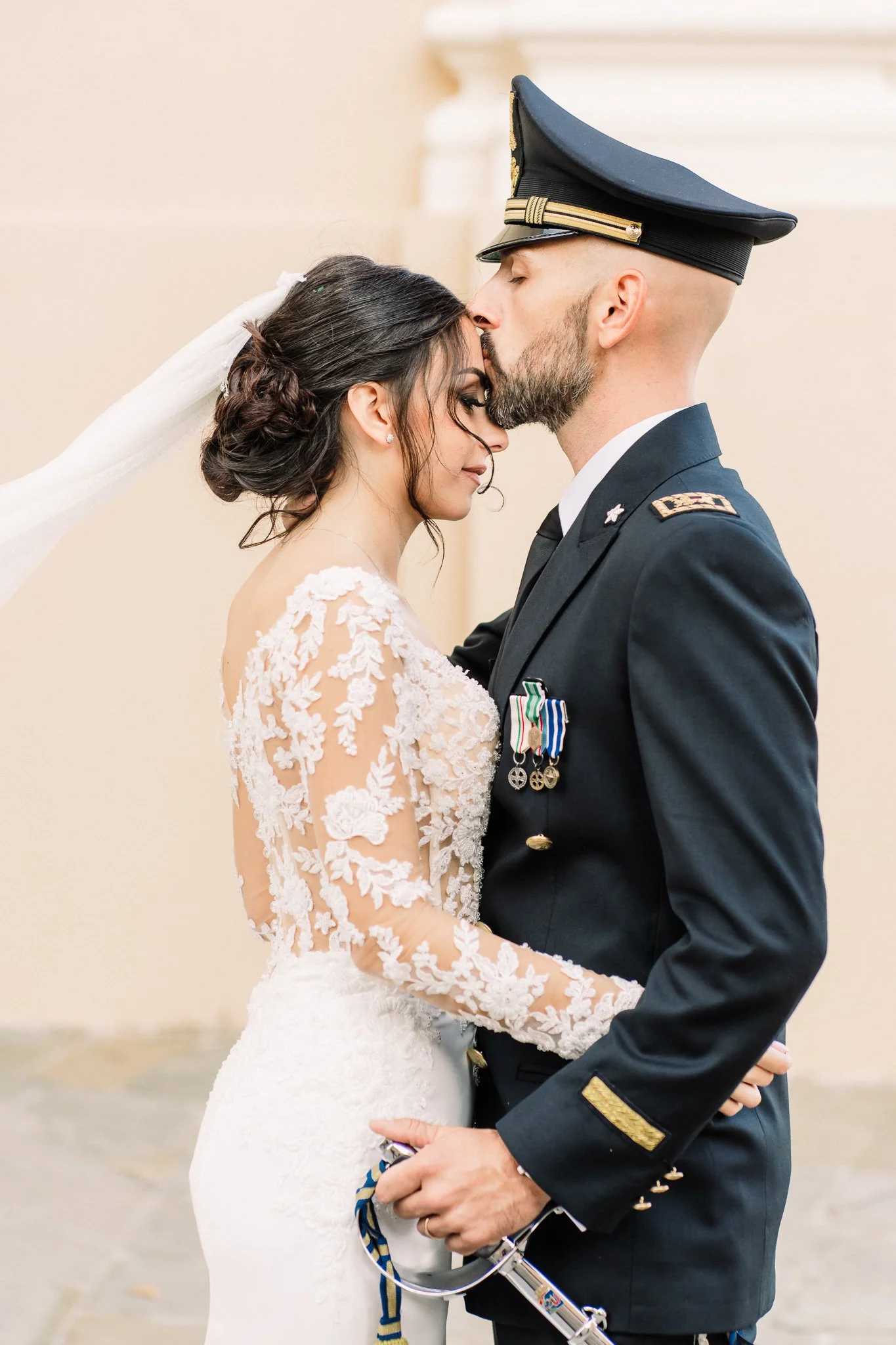 A bride and groom sharing a kiss, with the groom dressed in a U.S. military uniform and the bride in a lace wedding dress, during their wedding ceremony.
