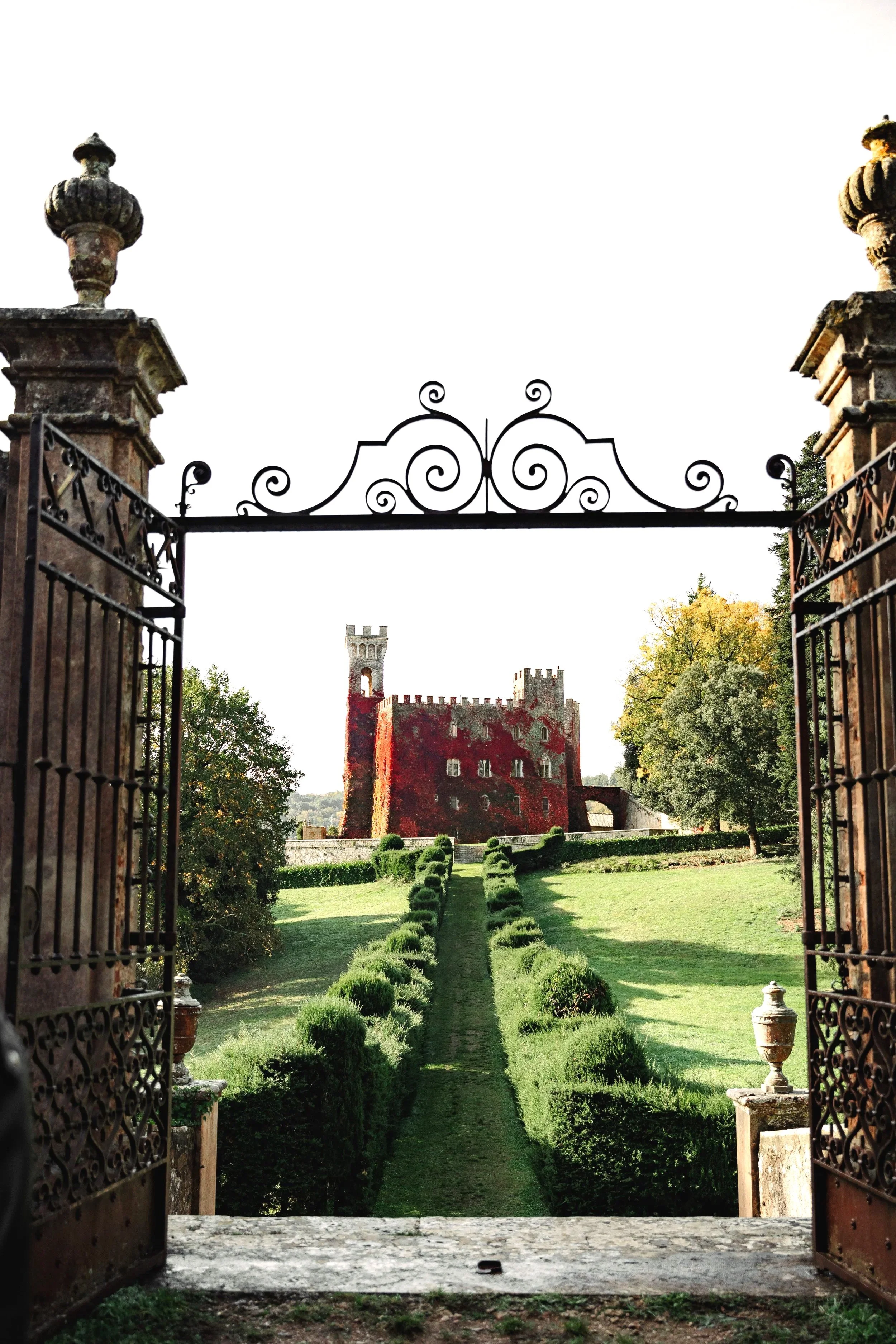  castle, Castello Di Celsa , luxury destination Wedding,viewed through an ornate iron gate with a stone pathway leading to it, surrounded by green grass and trees.