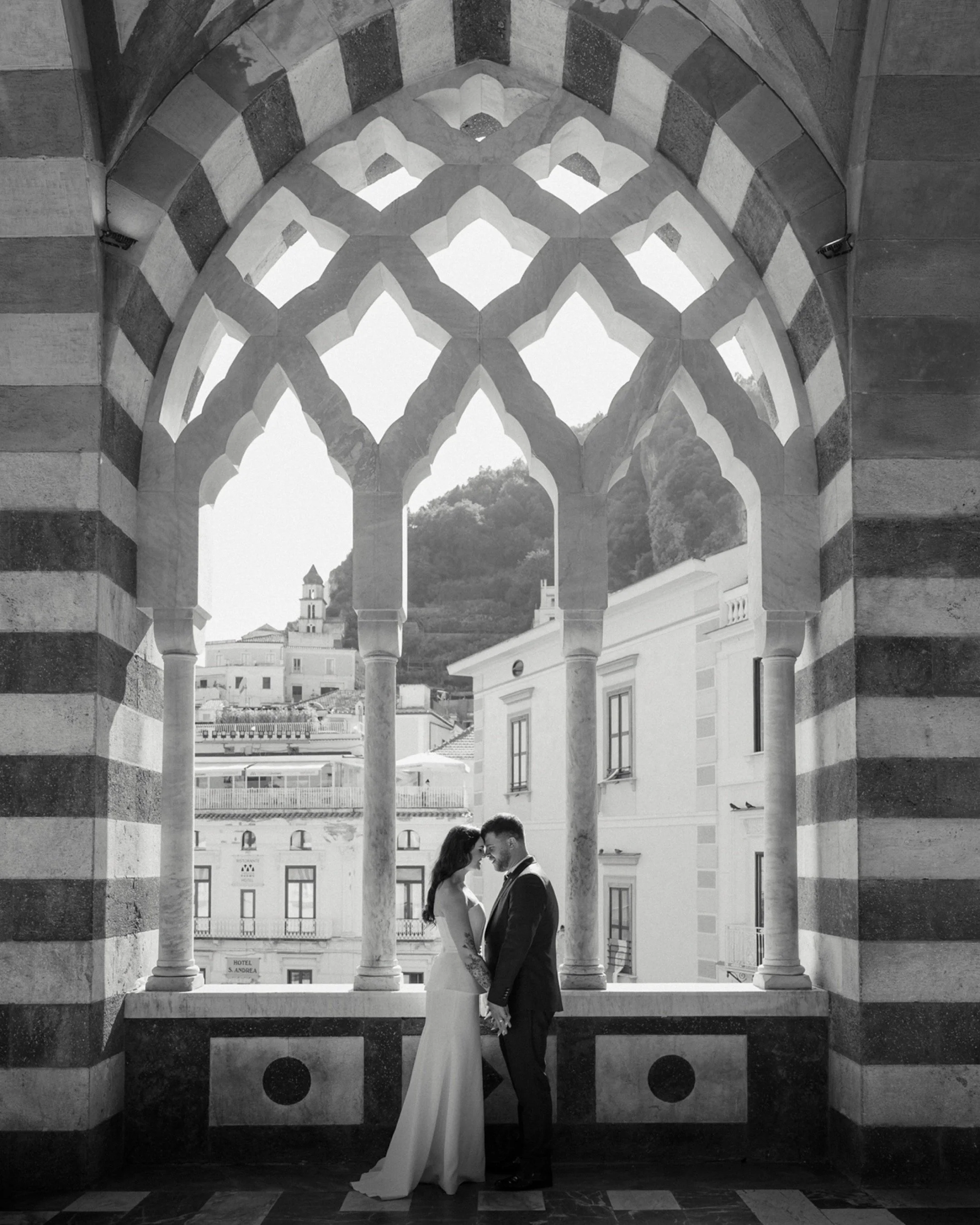 A bride and groom standing close together, holding hands, with foreheads touching, in front of an ornate arched window overlooking a cityscape with mountains in the background.