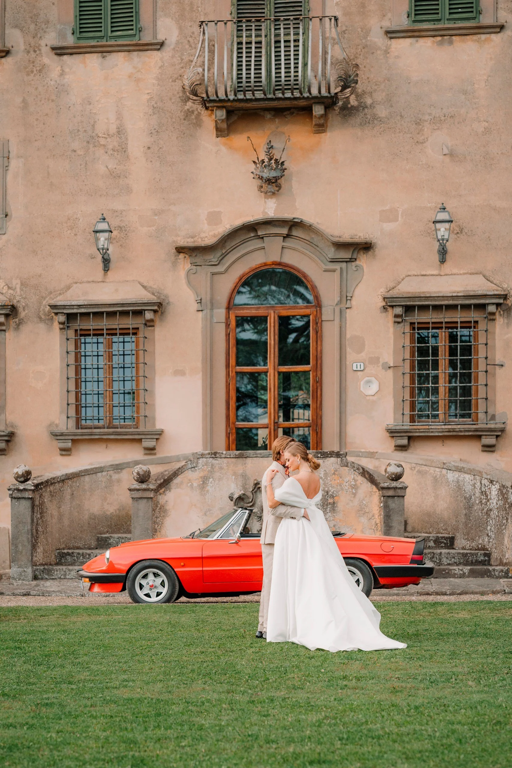 A bride and groom sharing a kiss in front of a vintage red convertible car with an old brick building in the background.