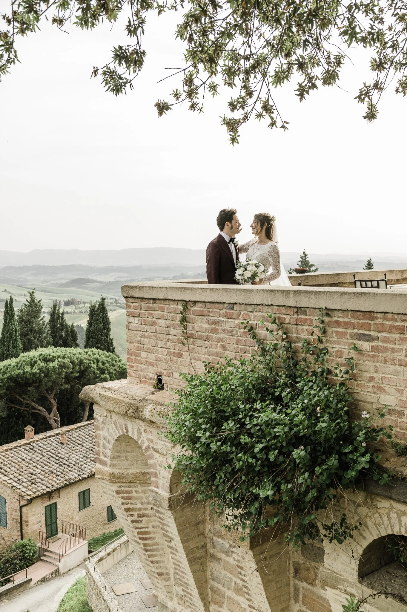 Bride and groom standing face to face on a brick terrace with a landscape view of hills and trees in the background.