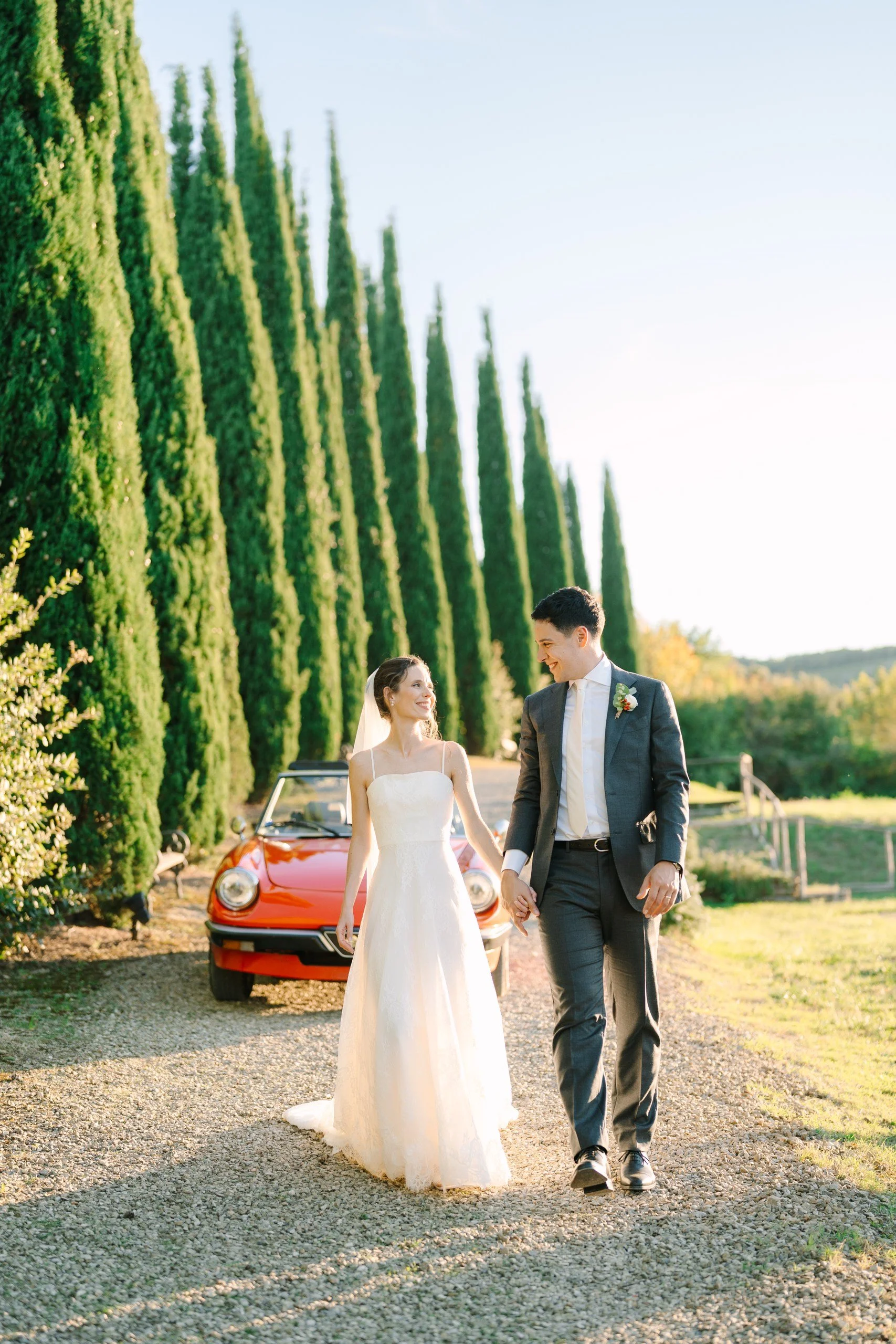 A newlywed couple walking hand in hand outdoors, with tall green trees and a red vintage car in the background, during late afternoon or early evening.