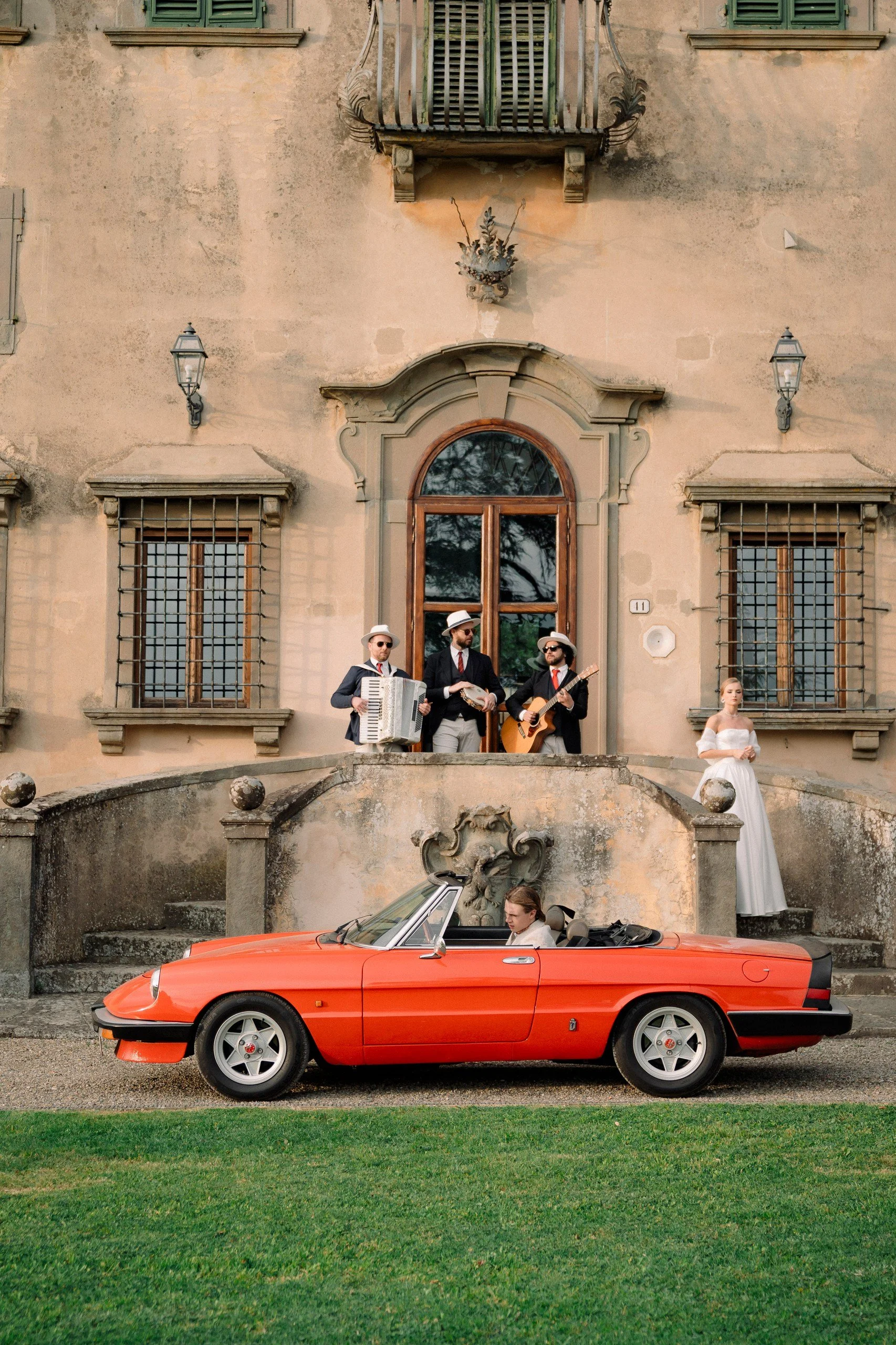 A vintage red convertible sports car parked in front of an old, ornate building with four soft-spoken musicians playing instruments on the steps above. A woman in a white dress stands to the right, looking away.