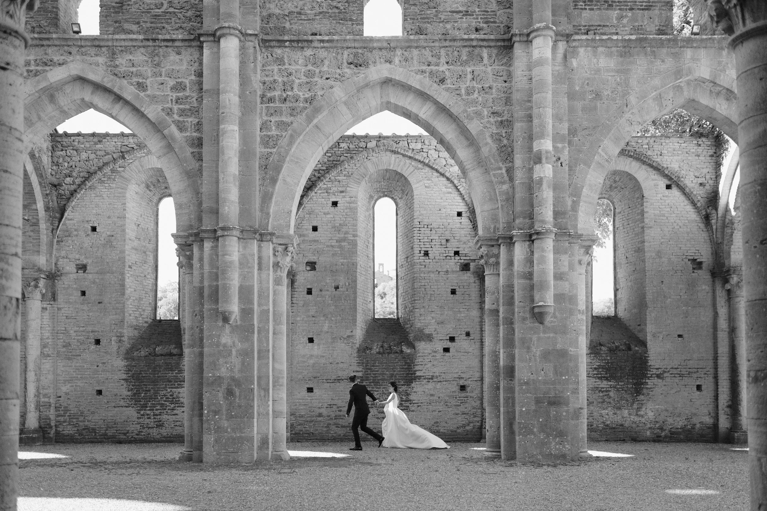 A wedding couple holding hands and dancing inside an old, partially ruined stone church with tall arches and large windows.