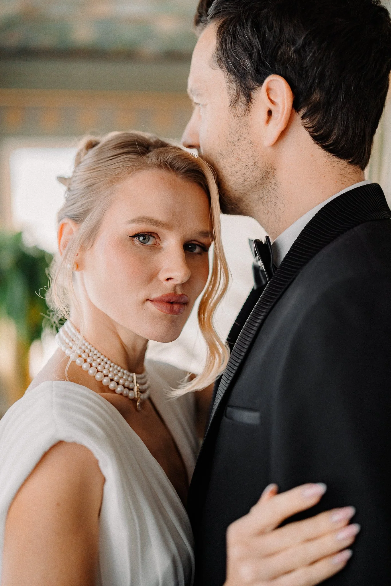 A close-up of a bride with blonde hair and blue eyes resting her hand on the chest of a groom with dark hair, dressed in a tuxedo, at a wedding. The bride wears a pearl necklace and a white wedding dress, looking directly at the camera; the groom is kissing her forehead, with a blurred background.