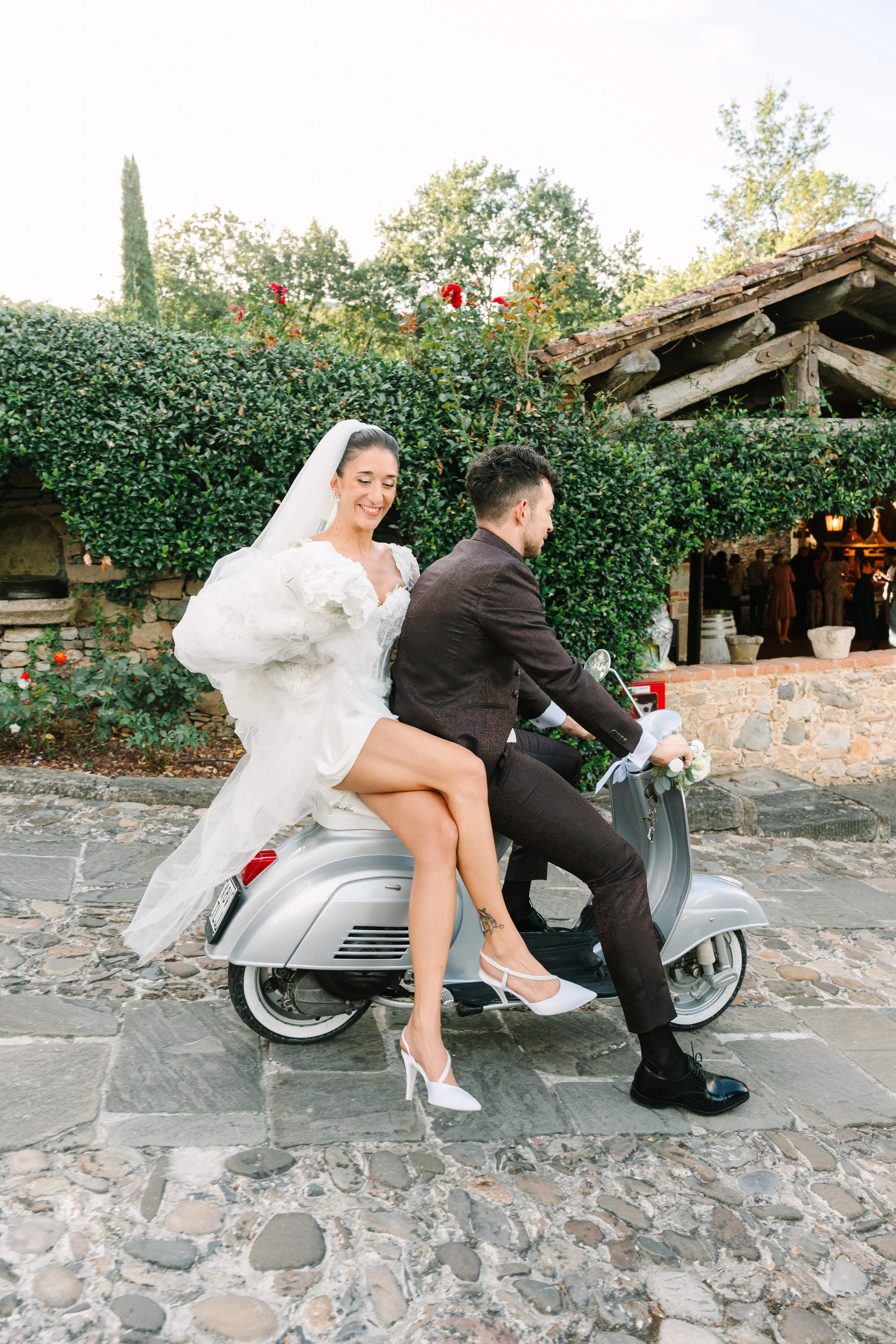 Bride in wedding dress and veil riding on a scooter with groom in formal suit, Valle di badia Sposa In vespa, matrimoni in toscana