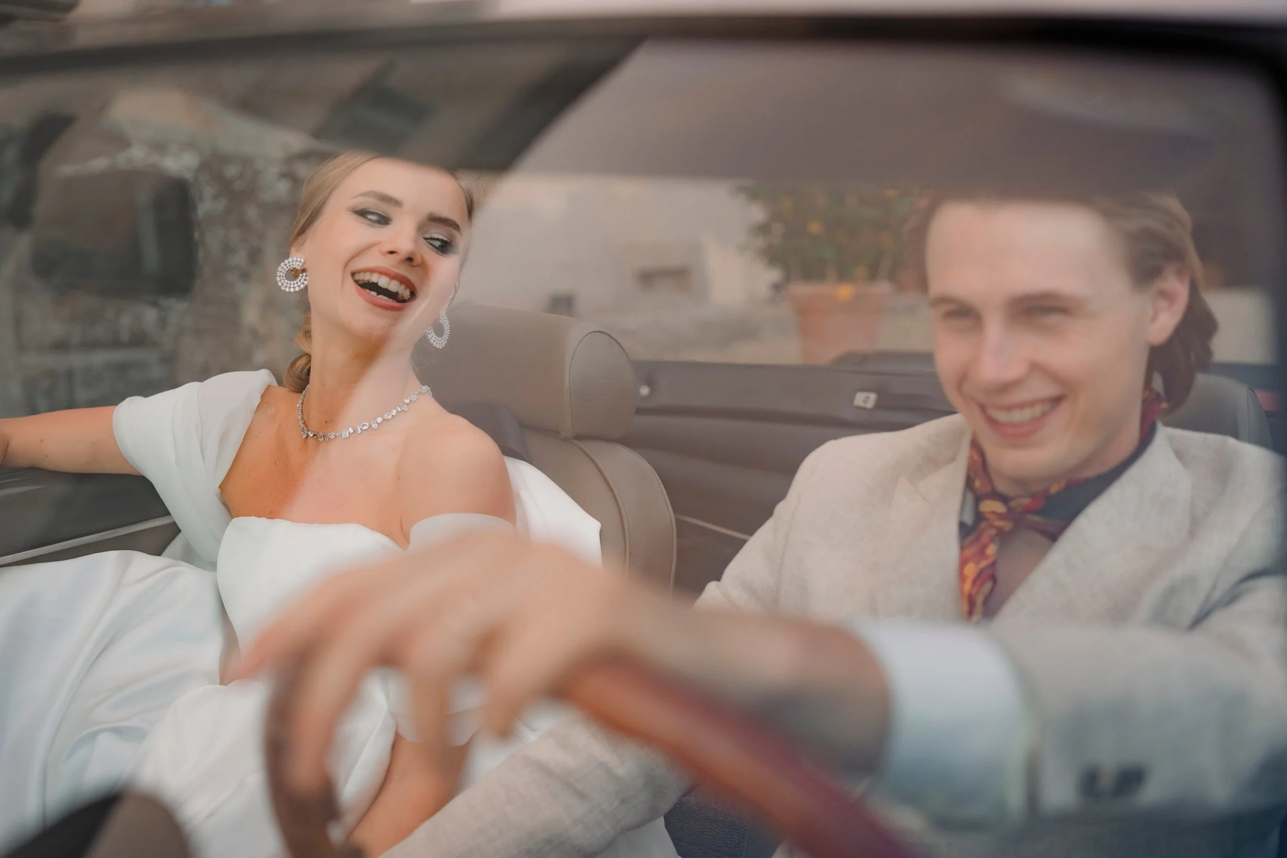 A woman in a white dress and jewelry laughs in a car with a man in a light-colored suit.