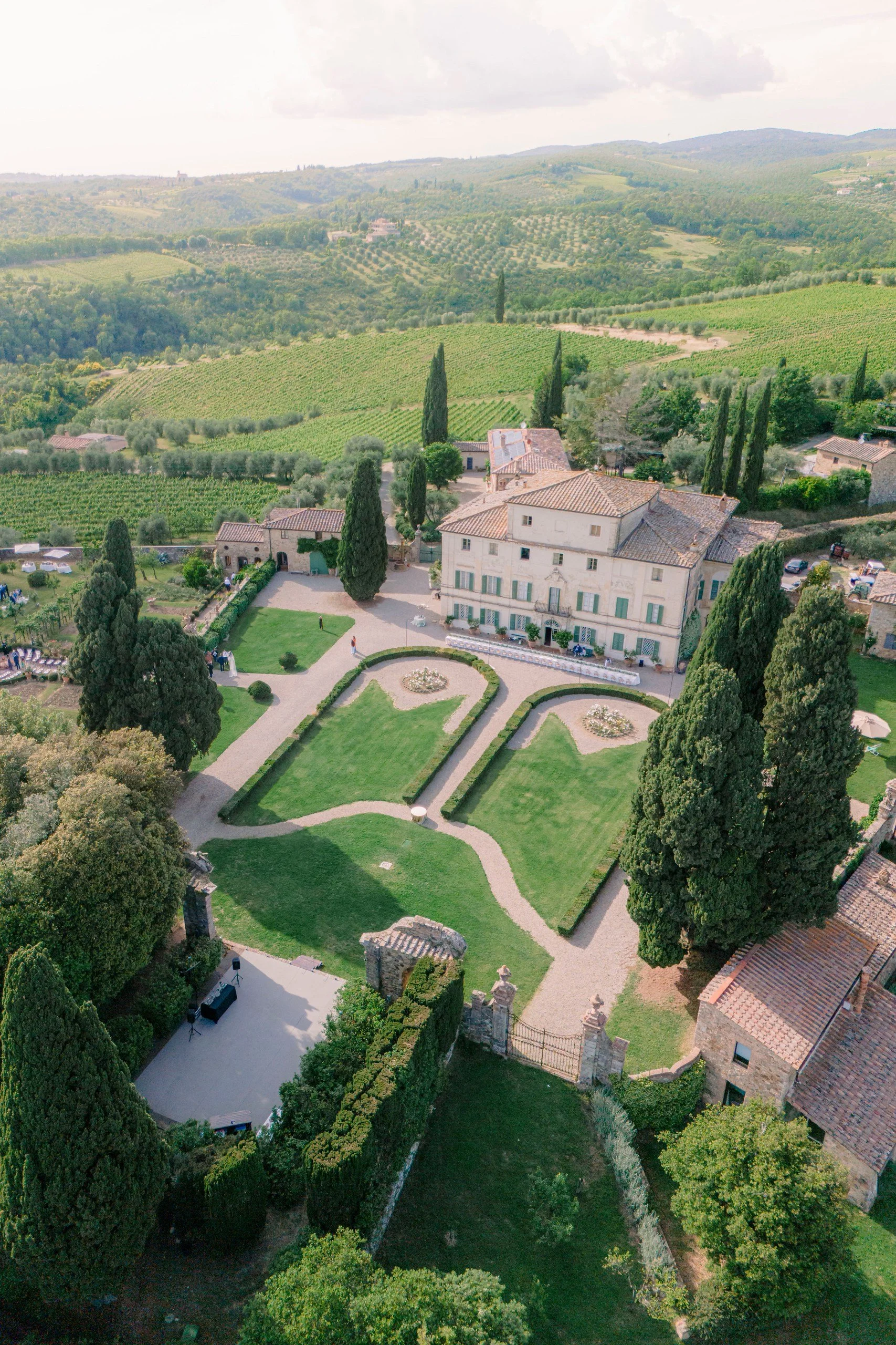 Aerial view of a large historic estate with well-manicured gardens and surrounding lush green landscape, rolling hills, and vineyards under cloudy sky.
