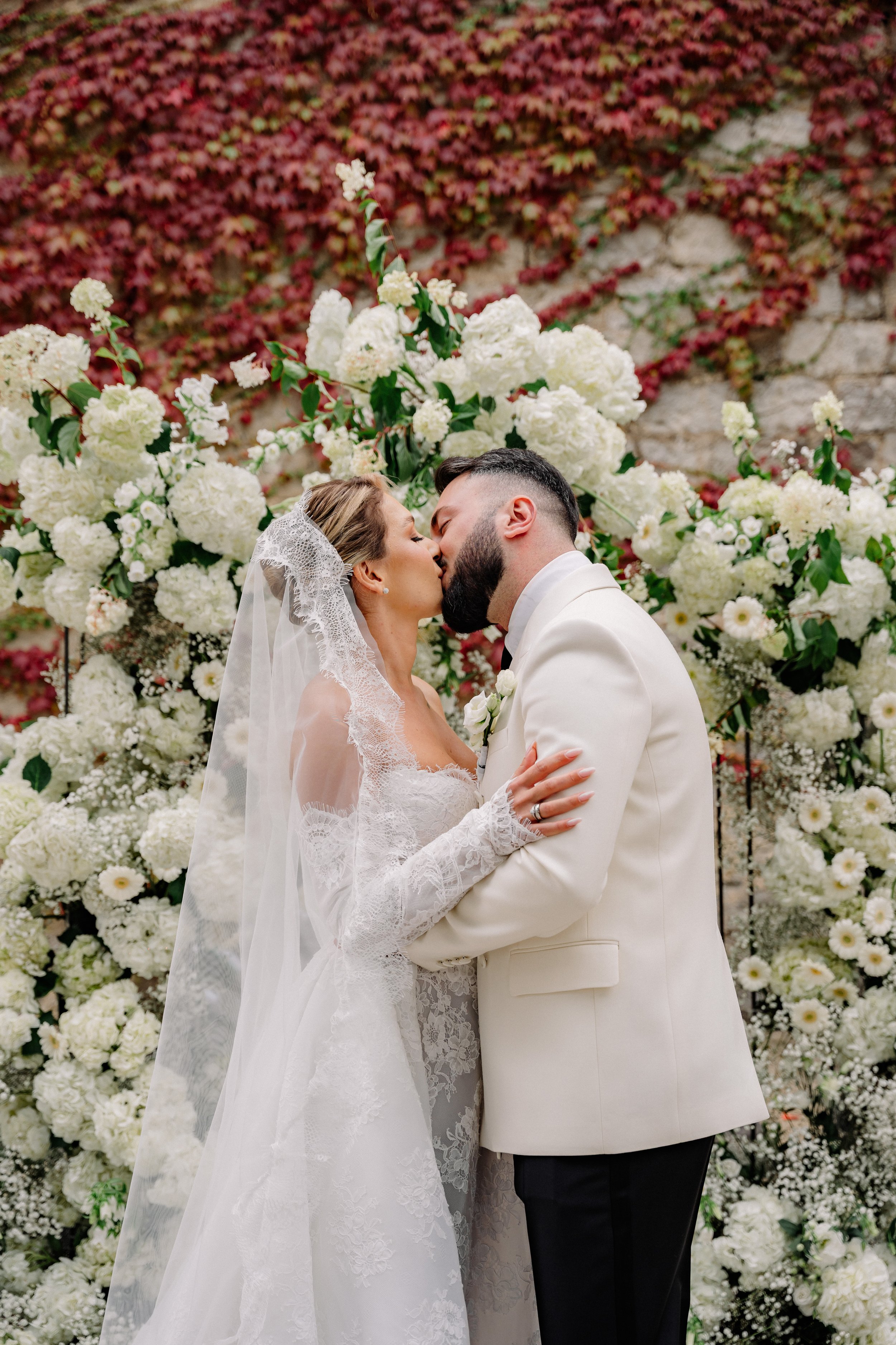 A bride and groom sharing a kiss in front of a floral backdrop of white flowers and greenery.