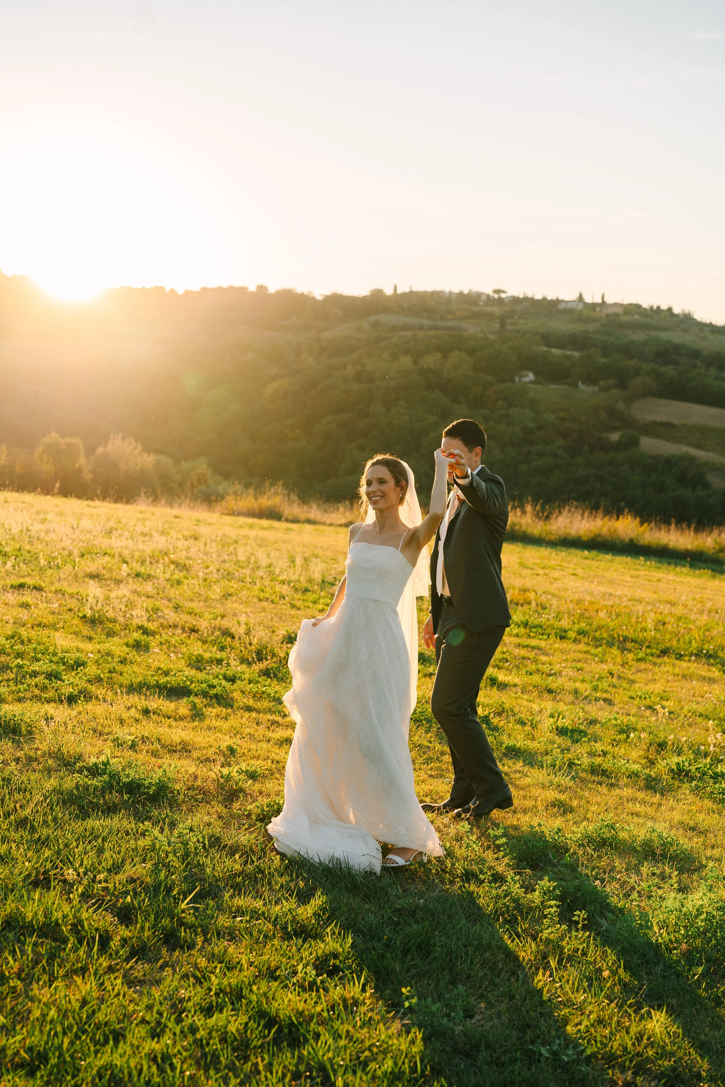 A bride and groom holding hands, dancing outdoors on a grassy hill at sunset, with hills and trees in the background.