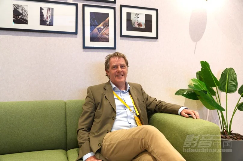 A man sitting on a green sofa in a room with framed pictures on the wall and a potted plant nearby.