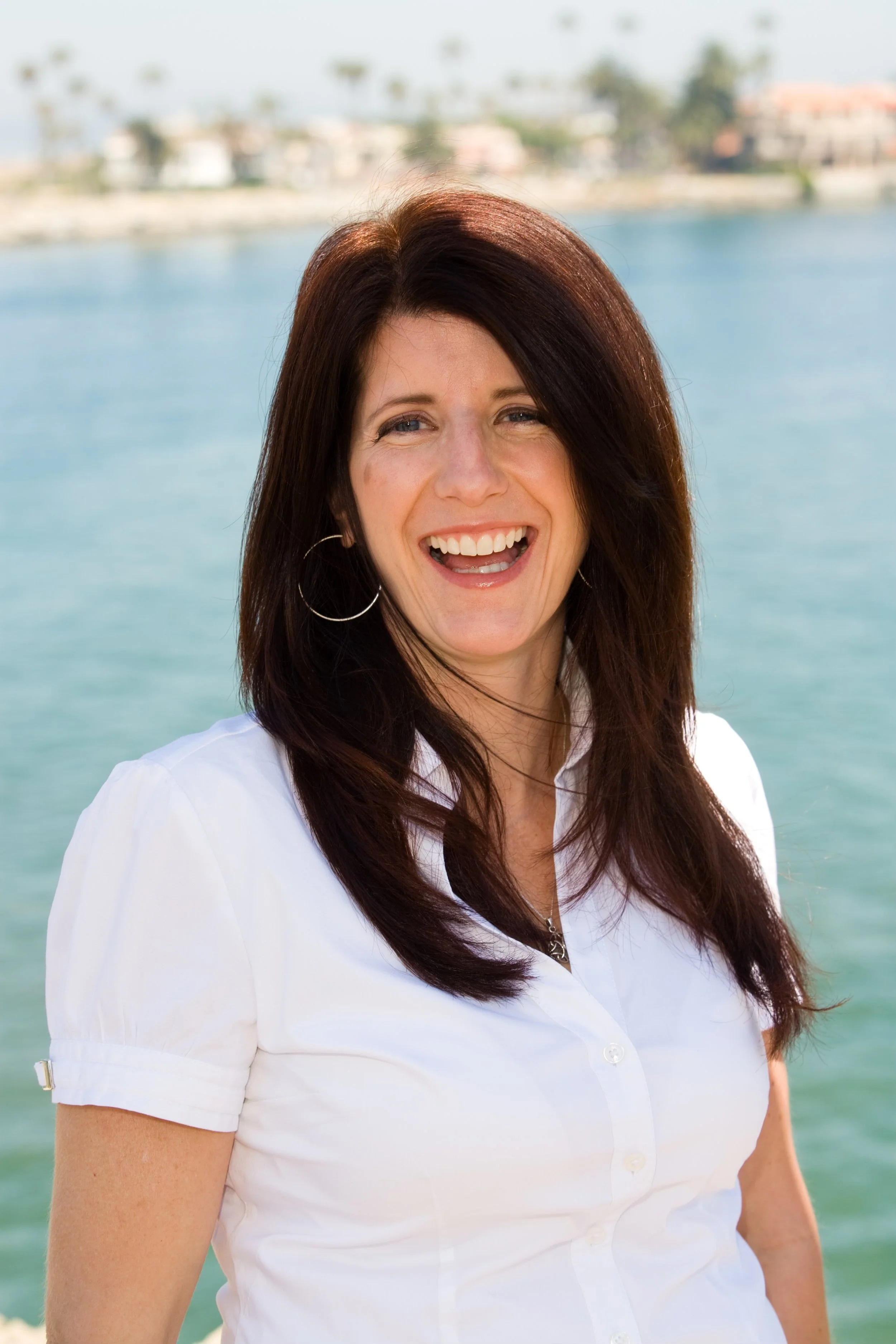 A woman with long brown hair wearing a white shirt, smiling outdoors with a body of water and buildings in the background.