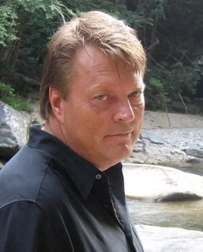 A man with light brown hair and a serious expression standing outdoors near a river with rocks and trees in the background.