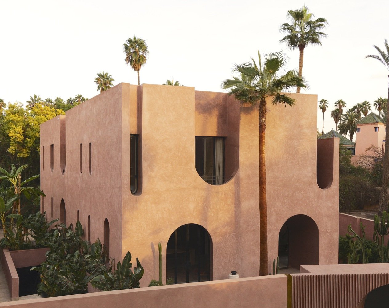 A modern pink stucco building surrounded by palm trees, with arched and elongated openings in a desert or warm climate setting.